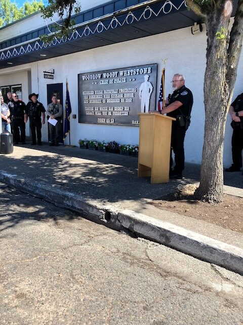 A Junction City police officer speaks from a podium during the Chief Woody Whetstone memorial dedication ceremony, with officers and honor flags standing alongside.