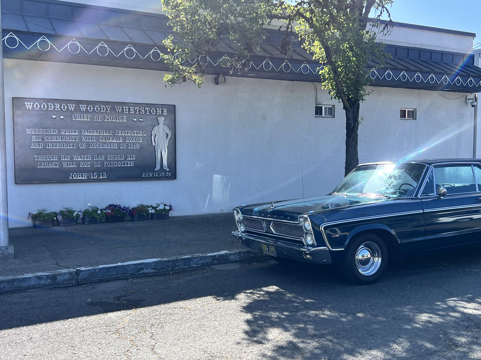 The Woody Whetstone memorial mounted on the wall of the Junction City Police Department, with flowers arranged below and a vintage patrol car parked at the curb.