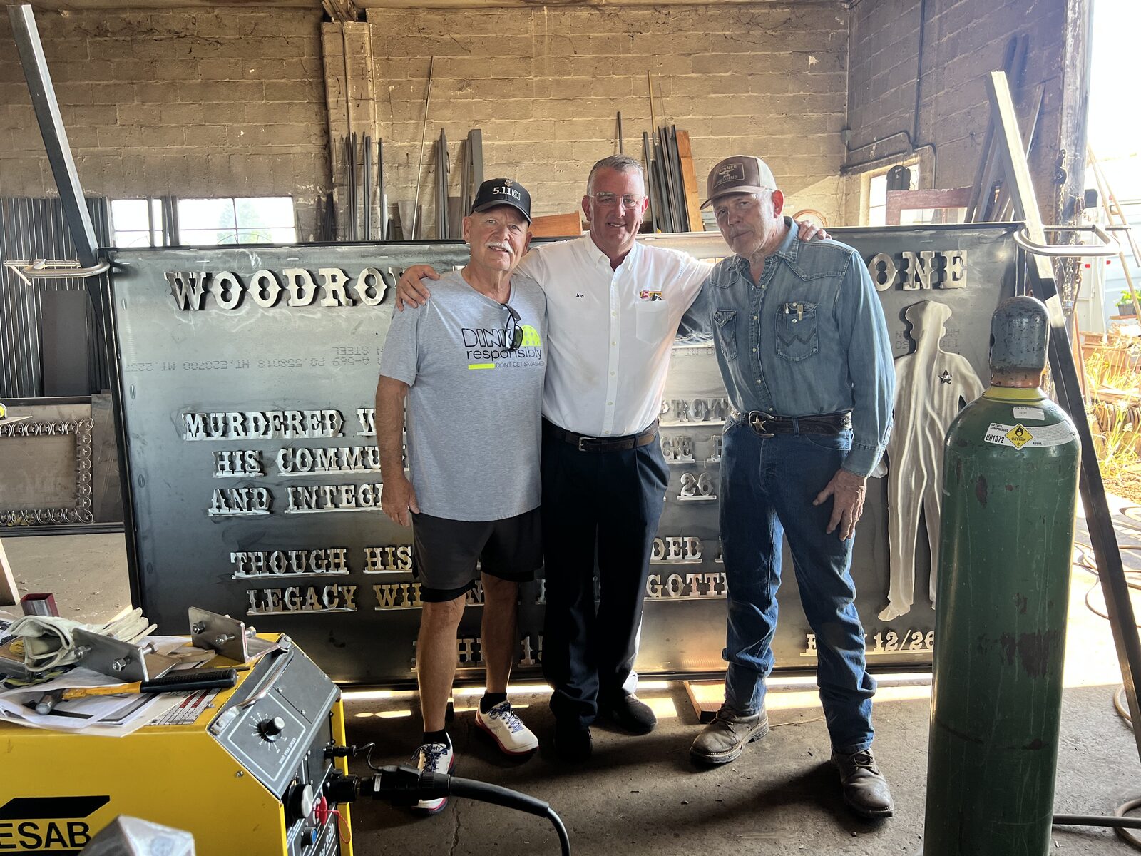 Three men stand with the finished Woody Whetstone memorial in the metal shop where it was fabricated, before installation in Junction City.